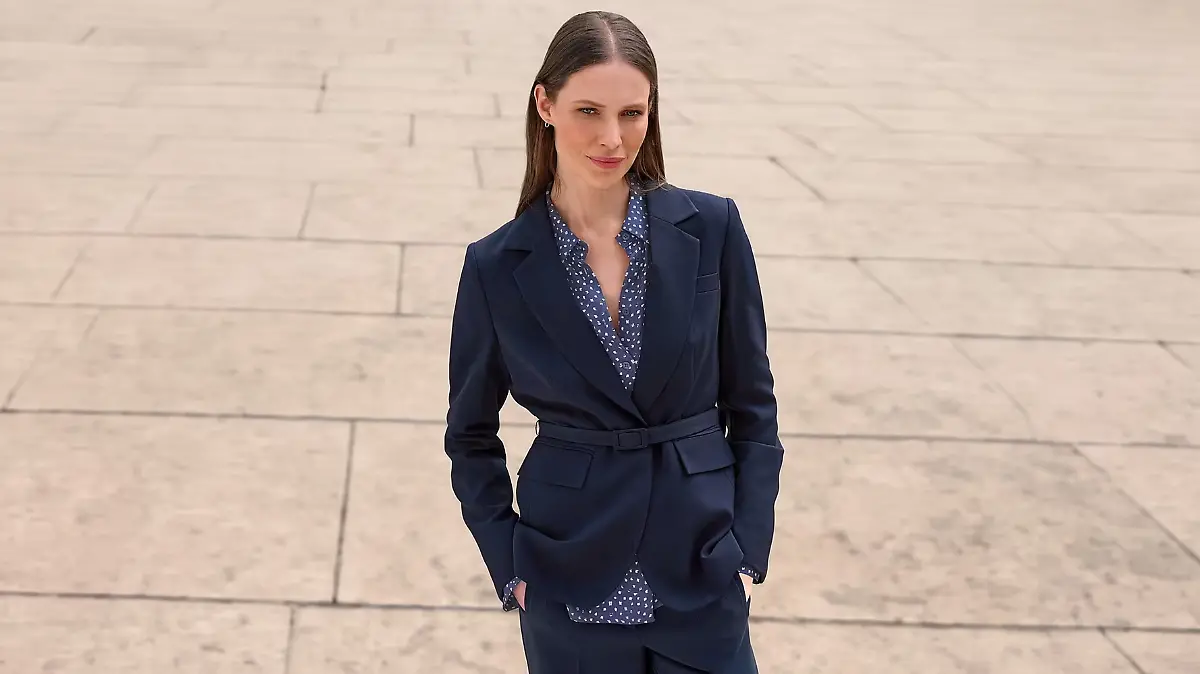 Model trägt einen Uta-Raasch-Blazer mit Taillengürtel, darunter eine gepunktete Bluse auf einem Gehweg.