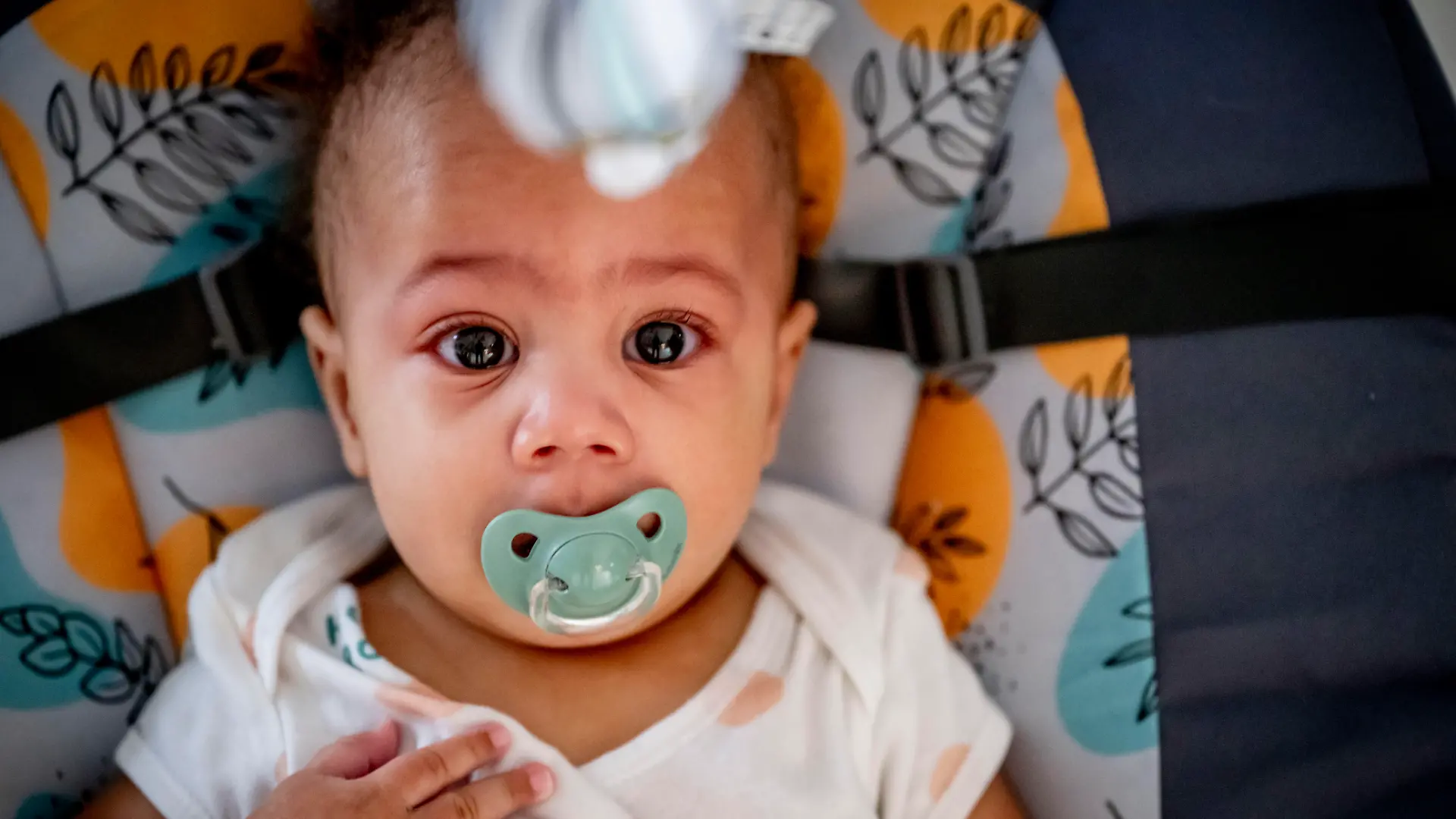 Baby mit Schnuller im Mund liegt in einer Trage und guckt in die Kamera.