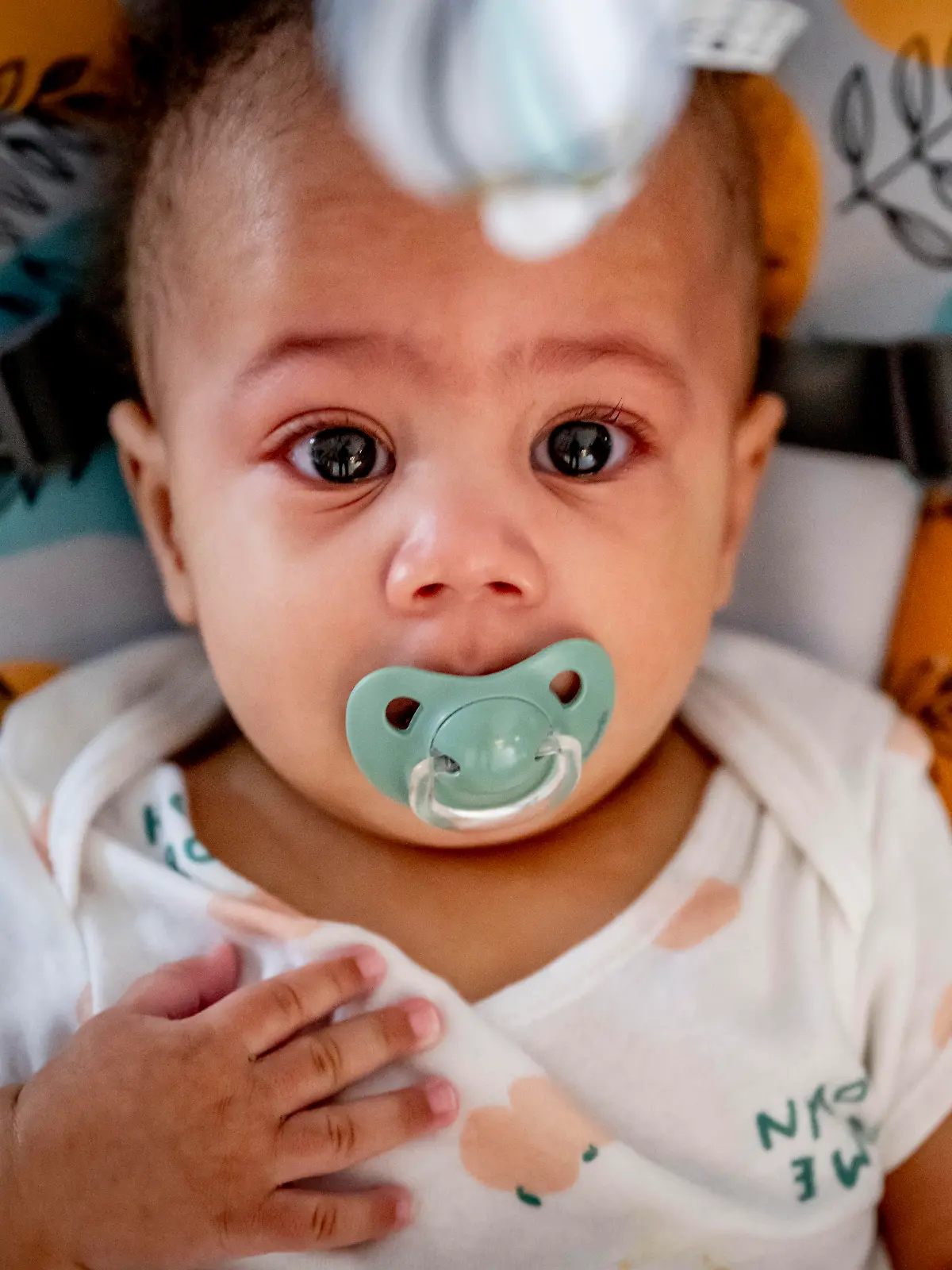 Baby mit Schnuller im Mund liegt in einer Trage und guckt in die Kamera.