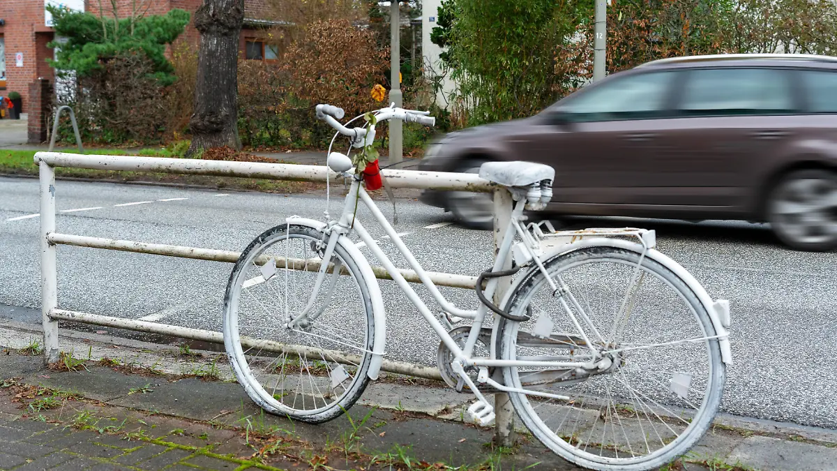 Das aufgestellte Ghostbike markiert eine Gefahrenstelle und erinnert an einen tödlichen Fahrradunfall. Ein schnell vorbeifahrendes Auto unterstreicht das Risiko, das Radfahrer durch rasenden Stadtverkehr erleben.