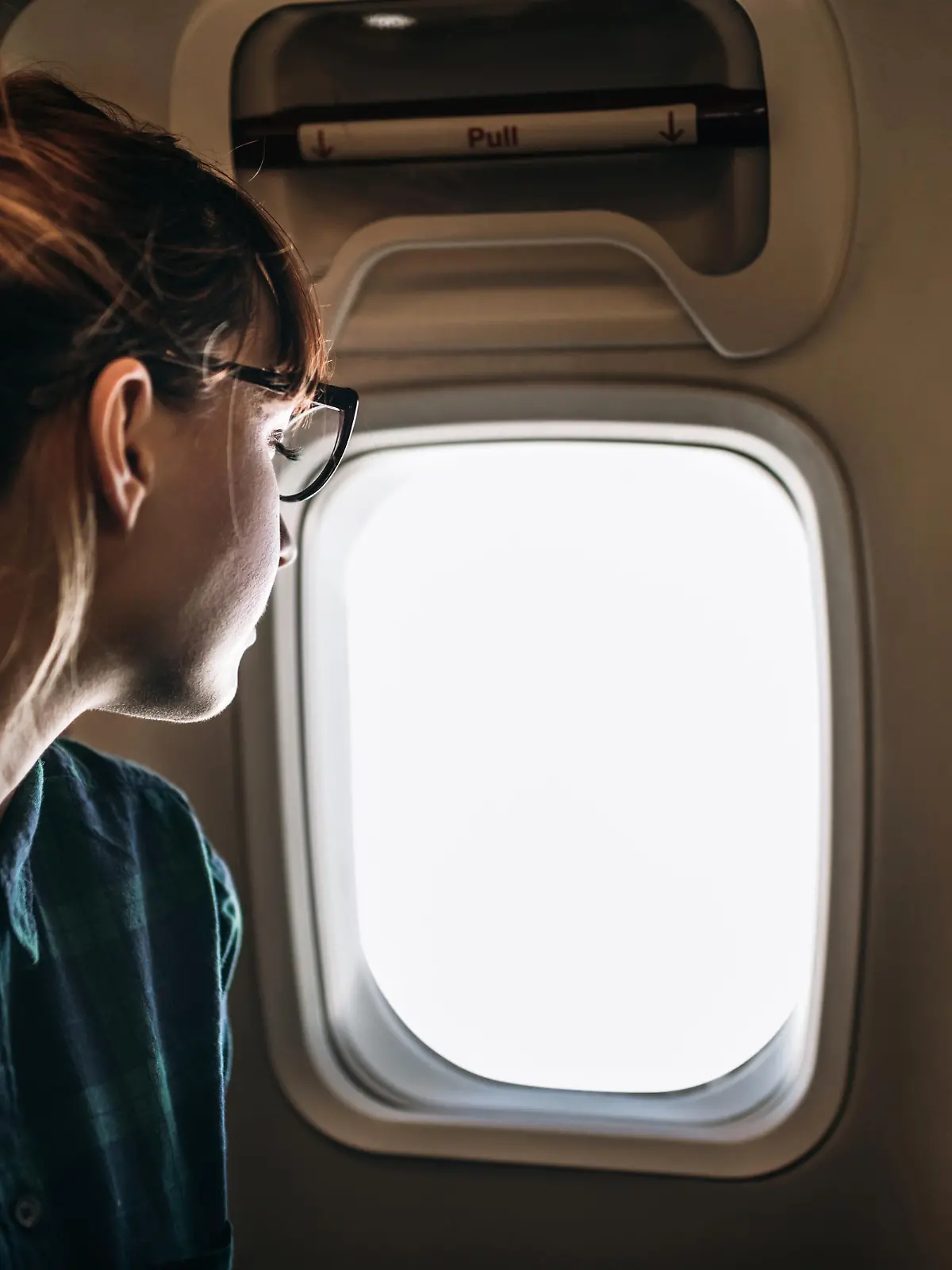 Eine junge Frau sitzt im Flugzeug auf einem Fensterplatz und schaut aus dem Fenster in den Himmel.