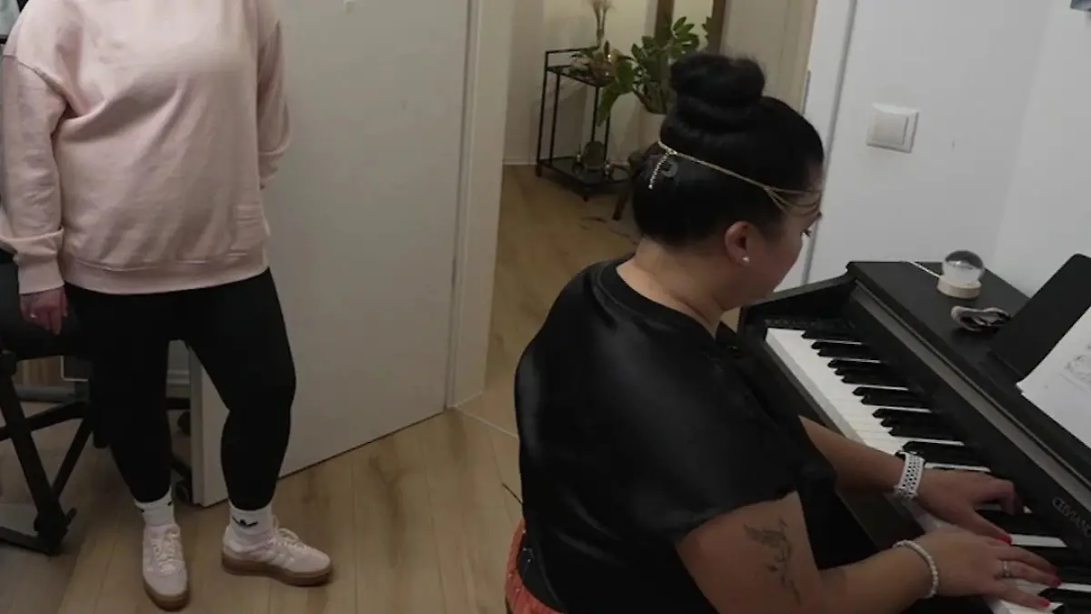 Das perfekte Dinner: Gastgeberin Pauline sitzt am Klavier, eine junge Frau steht daneben und schaut zu