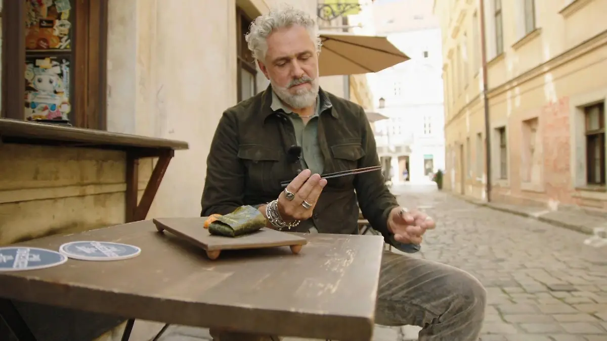 Roland schaut miesmutig auf Essstäbchen in seiner Hand, vor ihm eine Fischrolle.