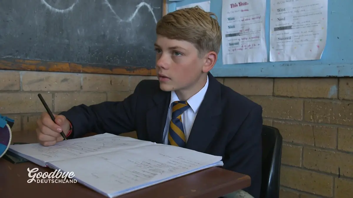 Goodbye Deutschland: Junge in dunkler Schuluniform mit weißem Hemd und gestreifter Krawatte sitzt vor einem aufgeschlagenen Heft an einem Schulpult