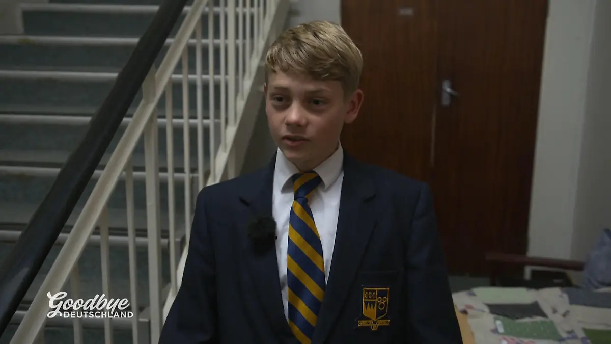Goodbye Deutschland: Junge in dunkler Schuluniform mit weißem Hemd und gestreifter Krawatte steht vor einer Treppe