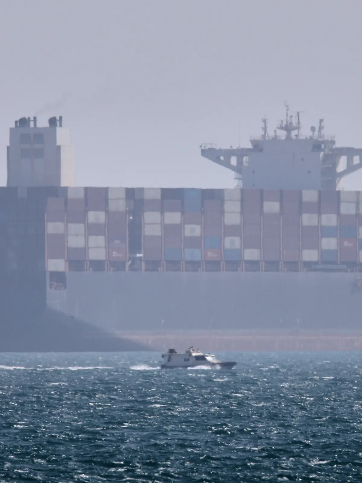 Ein Containerschiff liegt vor Anker, während im Vordergrund ein kleines Motorboot in der Straße von Hormus vor vorbeifährt. +++ dpa-Bildfunk +++
