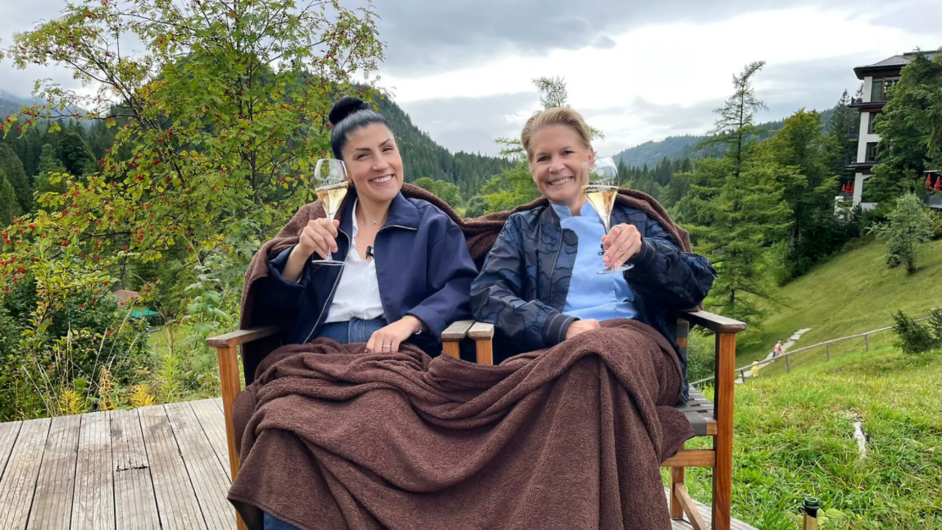 Elif Oskan (l.), Cornelia Poletto sitzen auf einer Bank im Bergpanorama und prosten mit Sekt in die Kamera