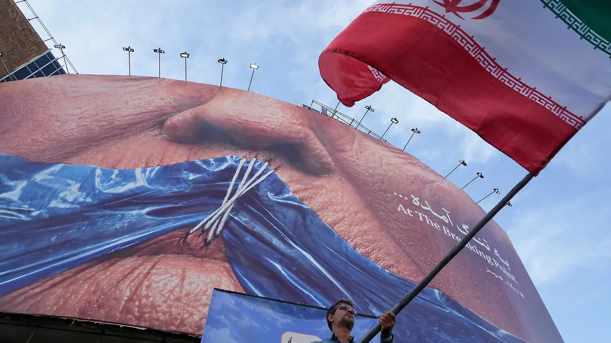 Ein Mann schwenkt bei einer regierungsnahen Aktion eine iranische Flagge vor einer Plakatwand mit einer Darstellung der Straße von Hormuz und zugenähten Lippen von US-Präsident Trump. +++ dpa-Bildfunk +++
