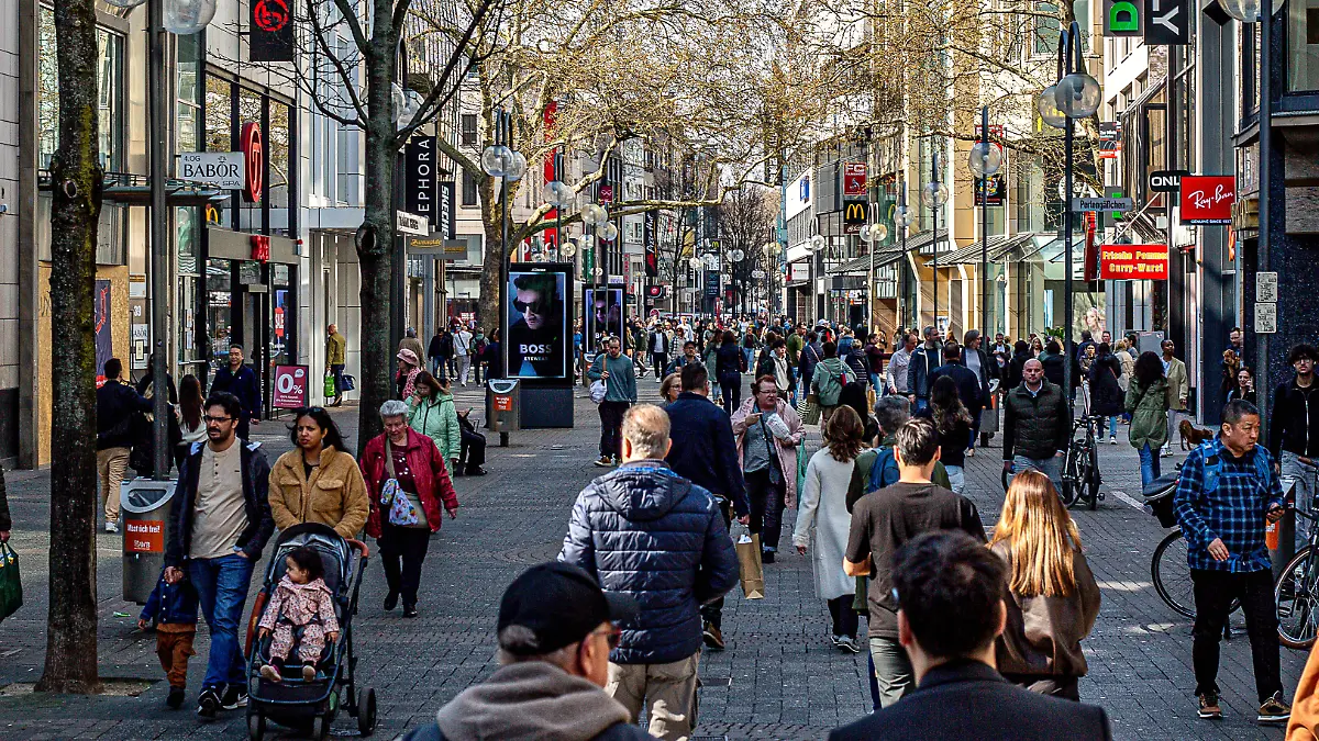 Diese Aufnahme zeigt das belebte Treiben von zahlreichen Passanten auf der sonnendurchfluteten Einkaufsmeile Schildergasse in Köln, gesäumt von markanten Ladenfronten und Bäumen.