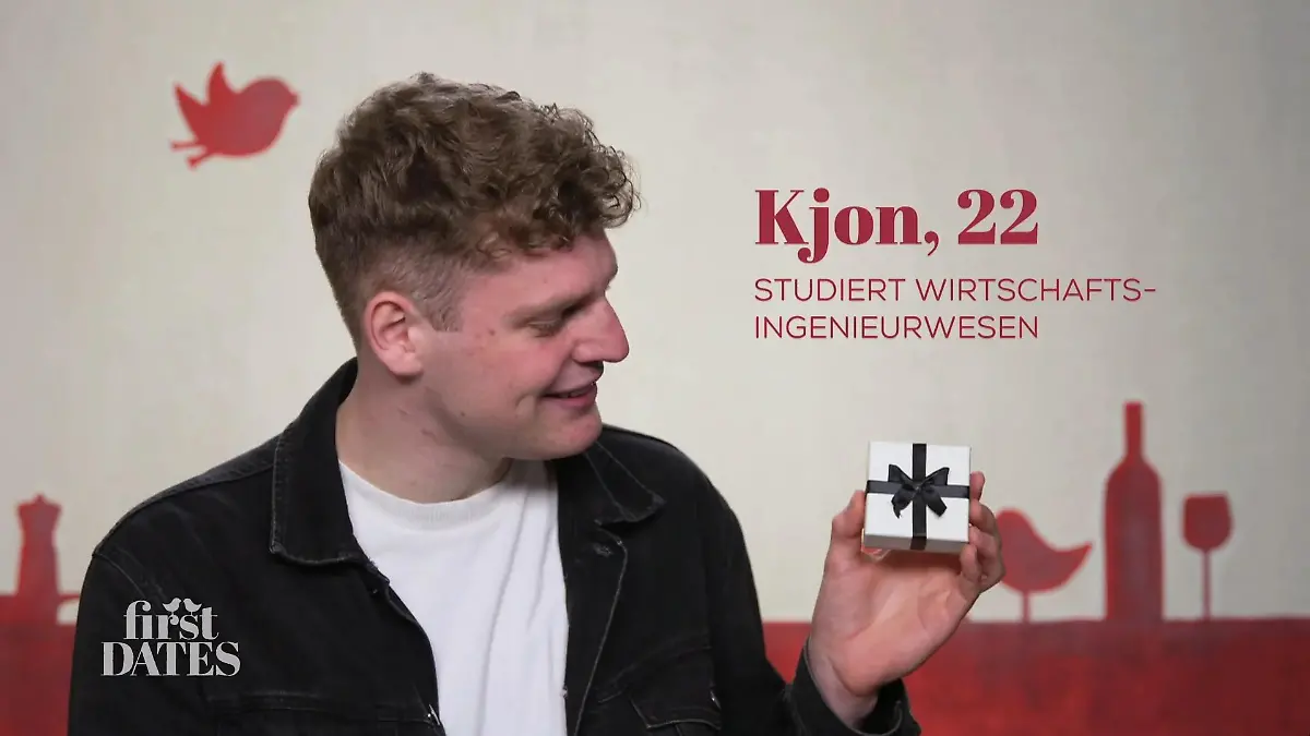 First Dates - Ein Tisch für Zwei: Junger Mann mit kurzen lockigen Haaren, hält eine kleine weiße Geschenkbox mit schwarzer Schleife in der Hand