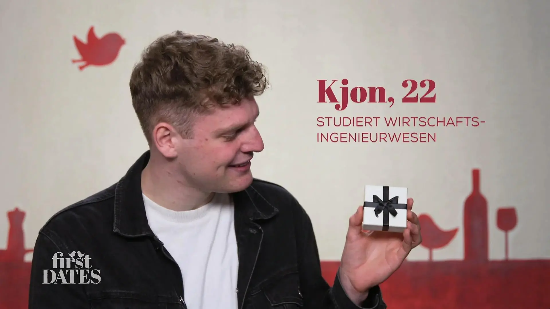First Dates - Ein Tisch für Zwei: Junger Mann mit kurzen lockigen Haaren, hält eine kleine weiße Geschenkbox mit schwarzer Schleife in der Hand