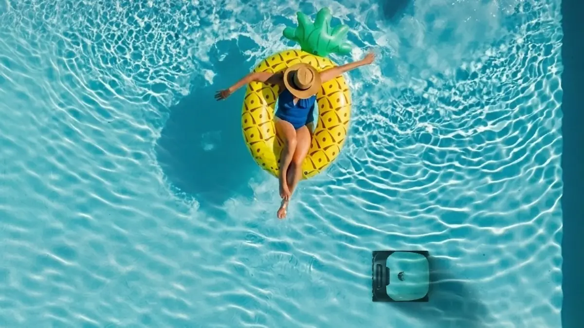 Frau mit Badeanzug und Sonnenhut planscht auf einem Schwimmring in Ananasform in einem Poolbecken. Unter ihr reinigt ein Poolroboter den Boden des Beckens.