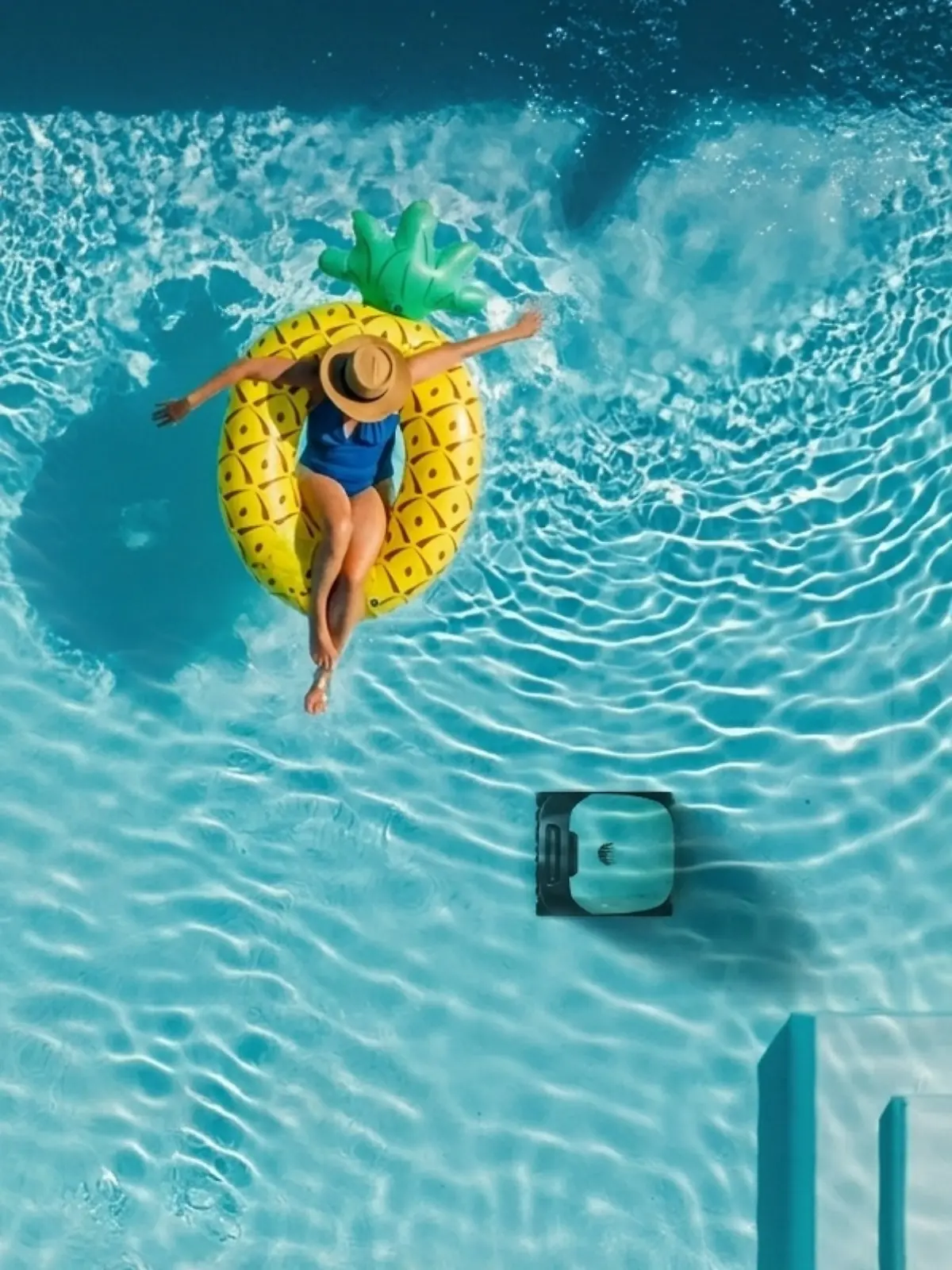 Frau mit Badeanzug und Sonnenhut planscht auf einem Schwimmring in Ananasform in einem Poolbecken. Unter ihr reinigt ein Poolroboter den Boden des Beckens.