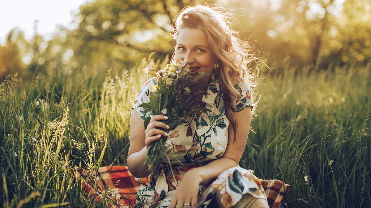 Frau in Blumenkleid sitzt auf einer Picknickdecke im Feld und riecht an einem selbstgepflückten Strauß.