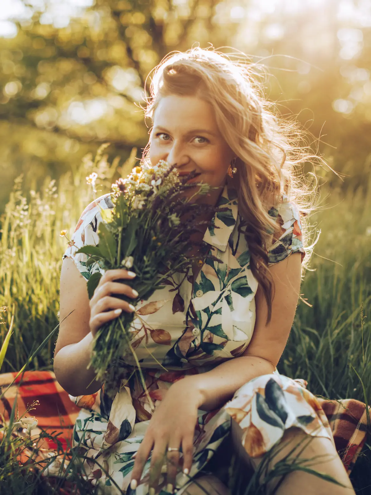 Frau in Blumenkleid sitzt auf einer Picknickdecke im Feld und riecht an einem selbstgepflückten Strauß.