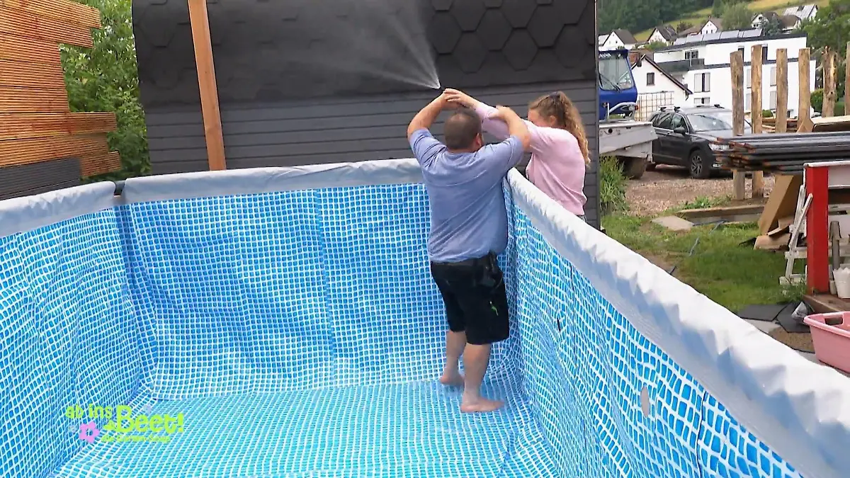 Ab ins Beet: Alex steht mit dem Schlauch außerhalb des Pools, Michel will ihn ihr aus der Hand nehmen