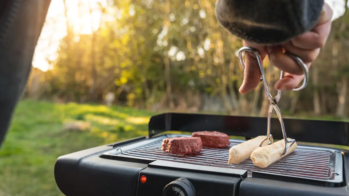 Junger Mann grillt im Garten bei schönem Wetter auf einem Elektrogrill. Auf dem Rost liegen vegane und vegetarische Fleischersatzprodukte wie Steak und Bratwurst als Alternative zu klassischem Grillgut