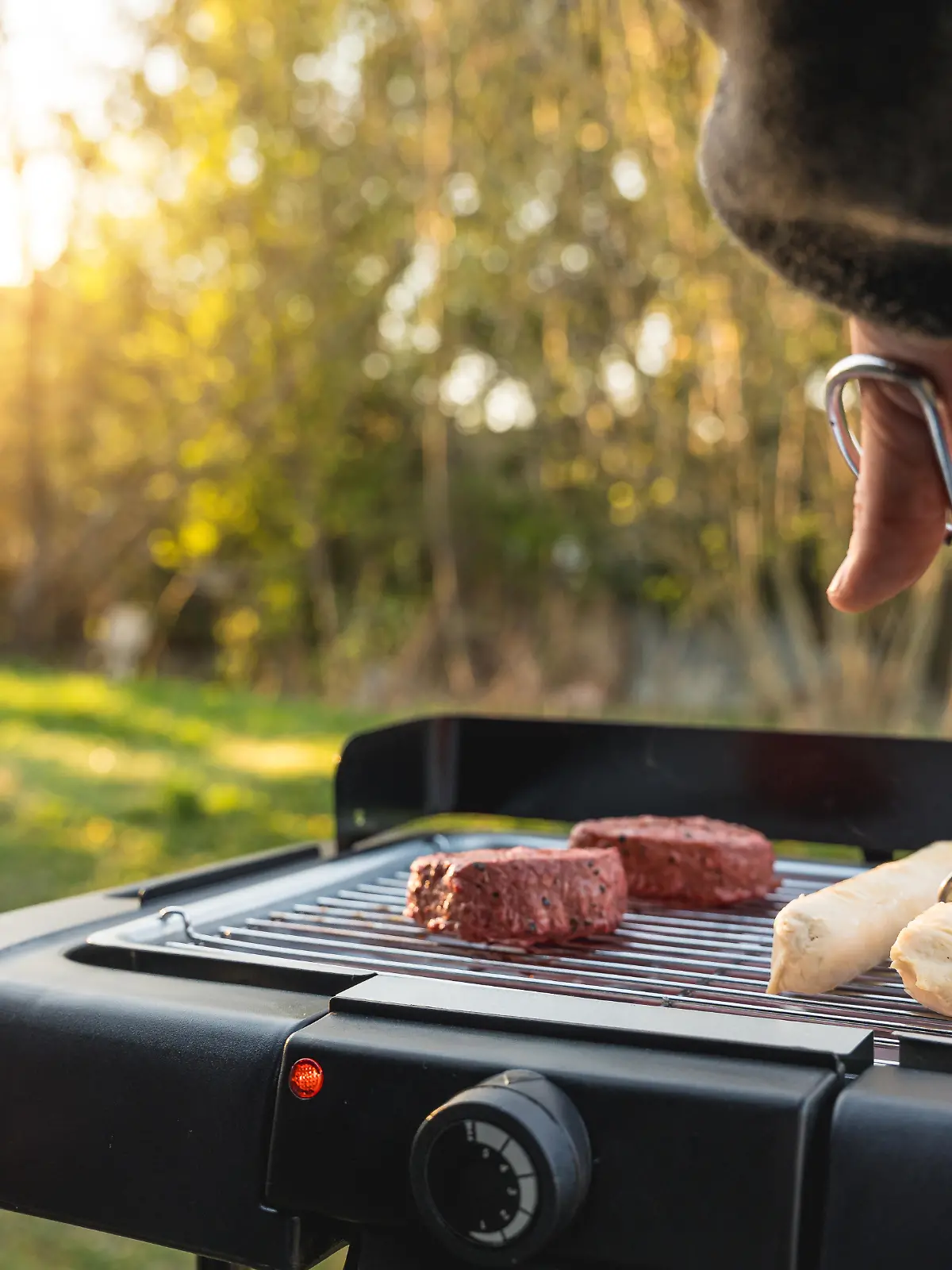 Junger Mann grillt im Garten bei schönem Wetter auf einem Elektrogrill. Auf dem Rost liegen vegane und vegetarische Fleischersatzprodukte wie Steak und Bratwurst als Alternative zu klassischem Grillgut