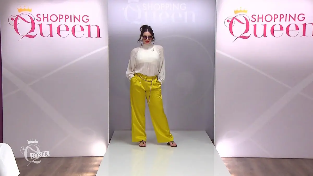Shopping Queen: Frau mit hochgesteckten Haaren, auffälliger Sonnenbrille, gelber, glänzender Hose und plissierter weißer, hochgeschlossener Bluse auf dem Catwalk