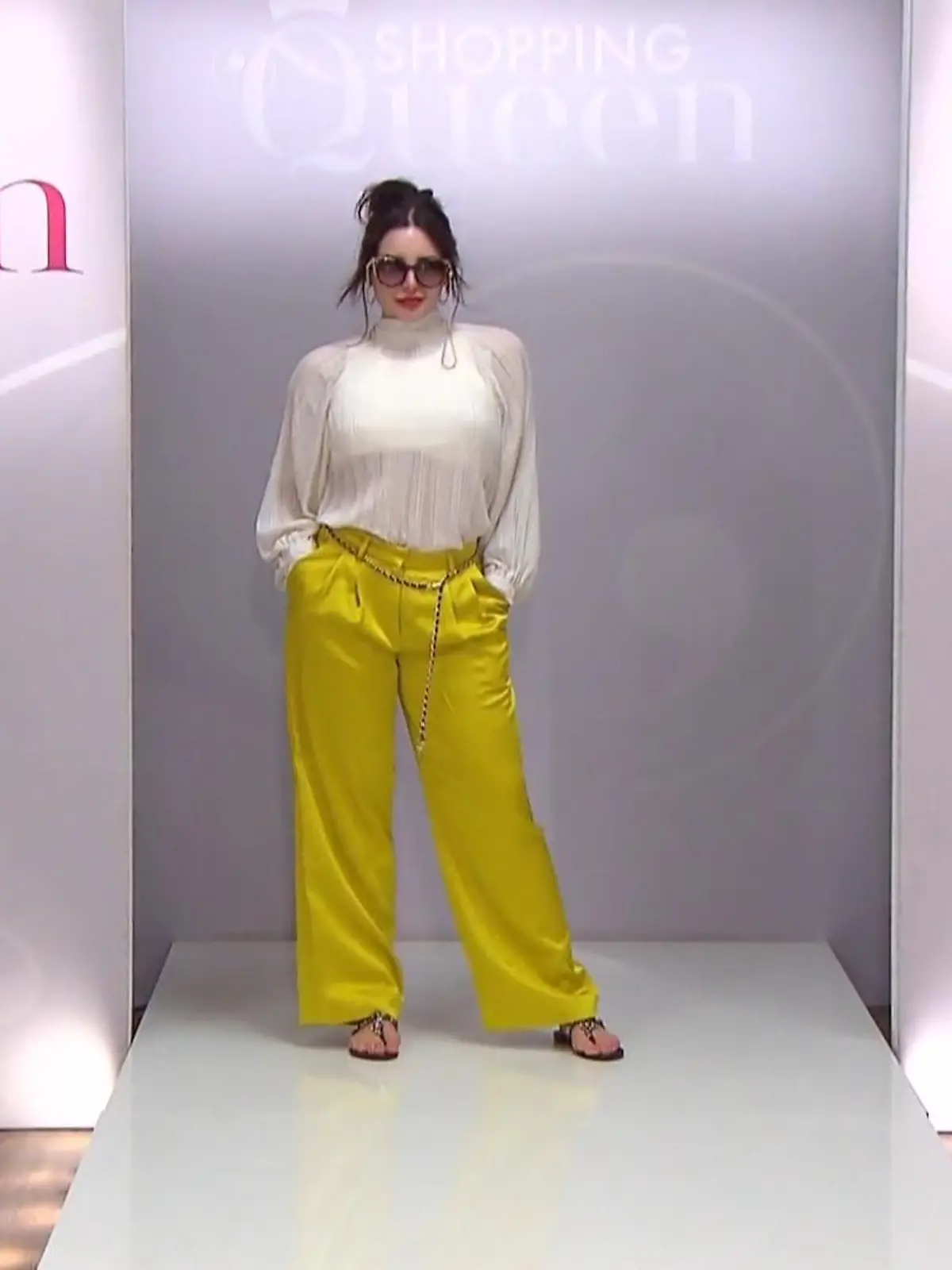 Shopping Queen: Frau mit hochgesteckten Haaren, auffälliger Sonnenbrille, gelber, glänzender Hose und plissierter weißer, hochgeschlossener Bluse auf dem Catwalk