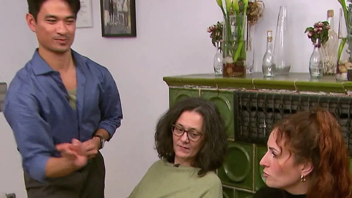 Ragil gestikuliert stehend am Tisch,  zwei Gäste sitzen lauschend daneben.