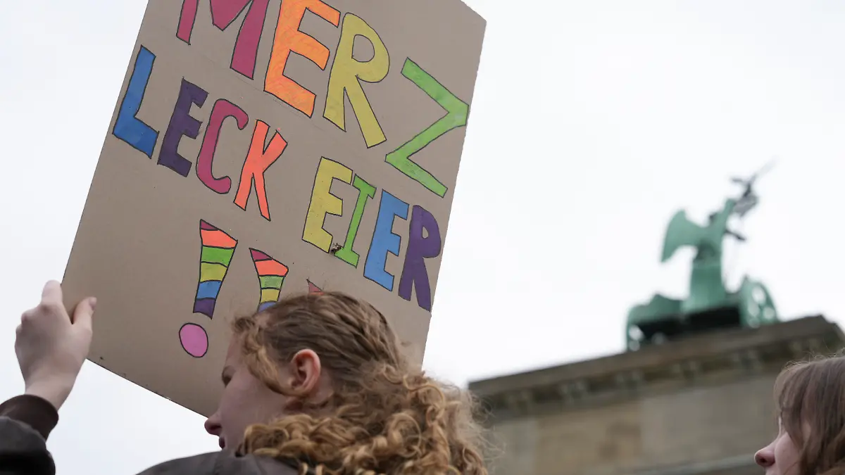 Eine Teilnehmerin einer Demonstration hält ein Plakat auf dem steht «Merz leck Eier!!!». Menschen versammeln sich vor dem Brandenburger Tor in Berlin um gegen die Wehrpflicht zu demonstrieren.