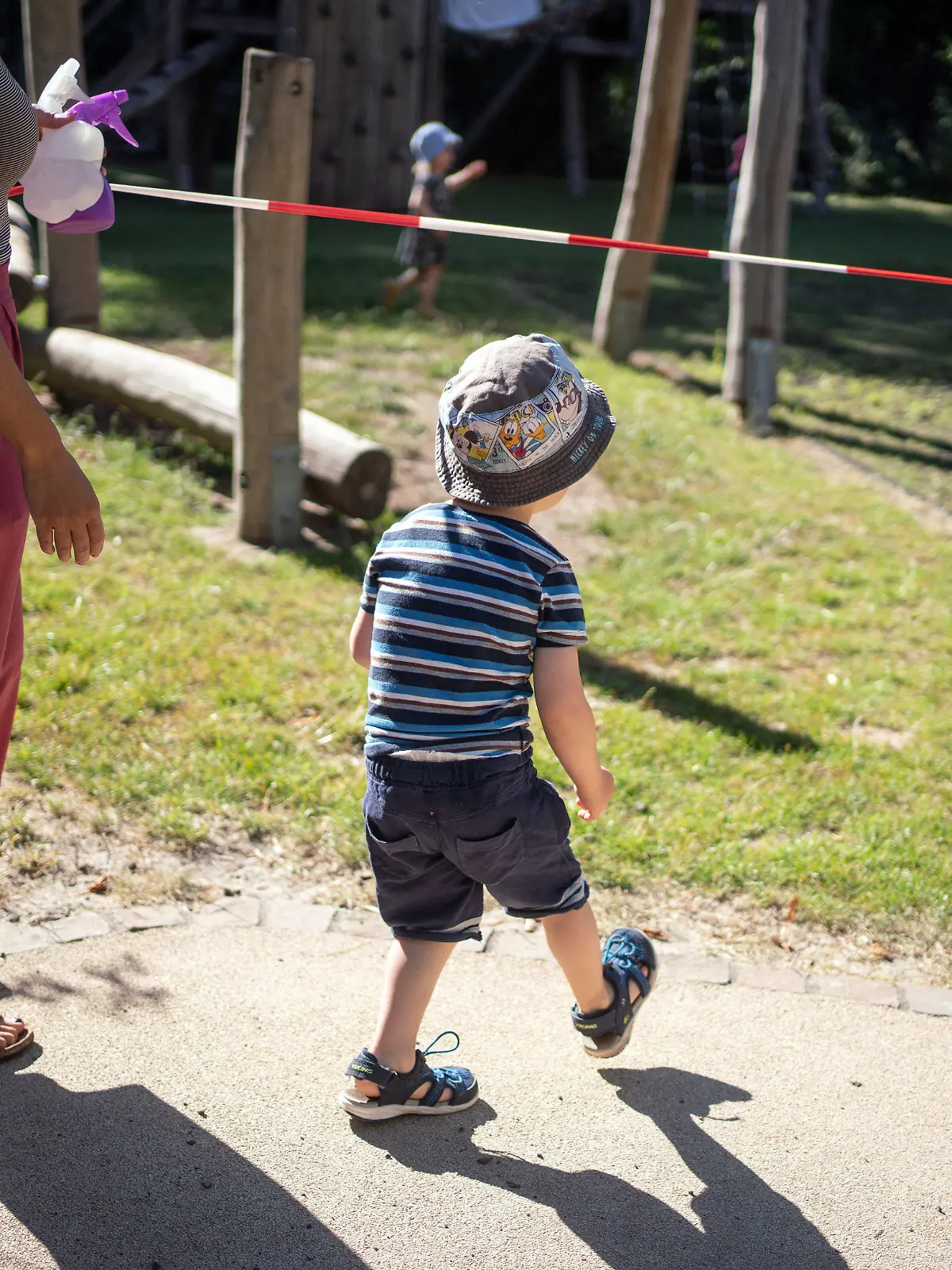 31.07.2020, Hessen, Frankfurt/Main: Ein Junge wird von einer Erzieherin (l) im Garten einer Kindertagesstätte im Stadtteil Bornheim unter einem Absperrband hindurch gelassen. Der Außenbereich ist in vier Teile abgegrenzt, in denen die Gruppen separat spielen können. Gewerkschaften erwarten einen schwierigen Start der Einrichtungen nach den Kita-Ferien in Hessen. Mit dem Ende der Sommerferien Mitte August müssen dann mehr Kinder als je zuvor in der Corona-Pandemie in den Kitas betreut werden. Foto: Frank Rumpenhorst/dpa +++ dpa-Bildfunk +++