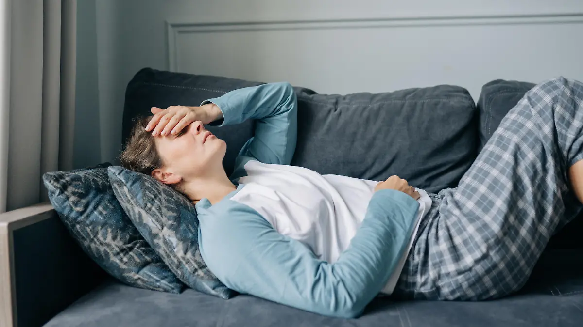 Frau liegt im Pyjama mit Migräne auf dem Sofa, hält sich die Hand übers Gesicht