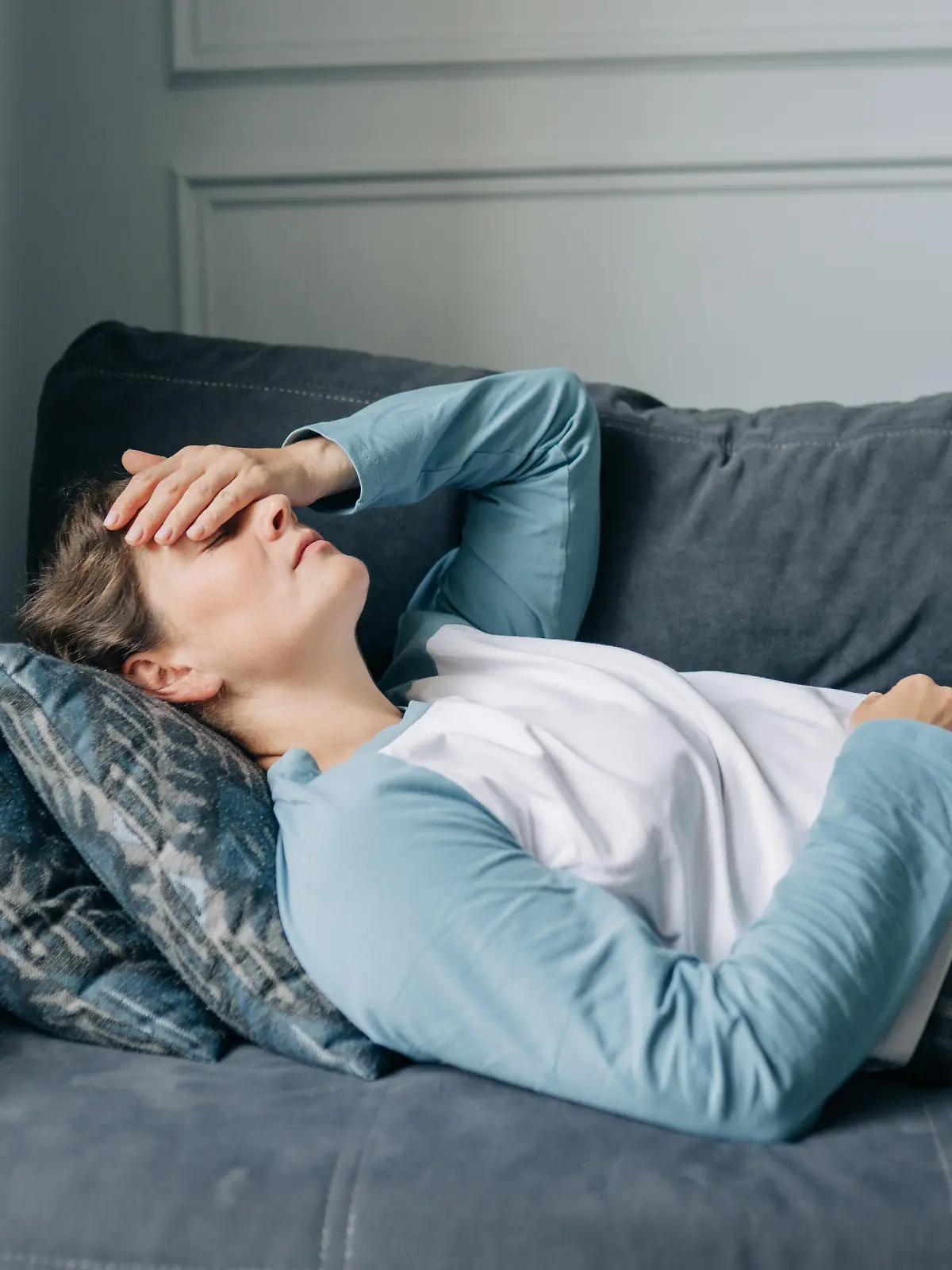 Frau liegt im Pyjama mit Migräne auf dem Sofa, hält sich die Hand übers Gesicht