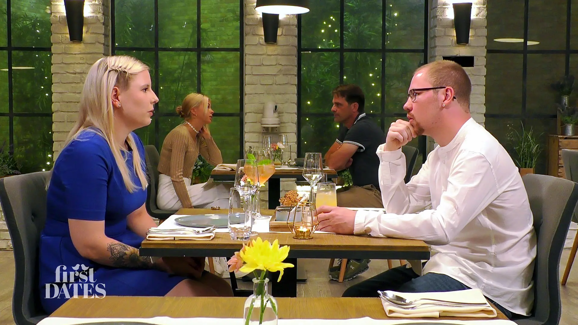 First Dates - Ein Tisch für zwei: Junge Frau im blauen Kleid und junger Mann im weißen legeren Hemd sitzen sich beim Date am Tisch gegenüber