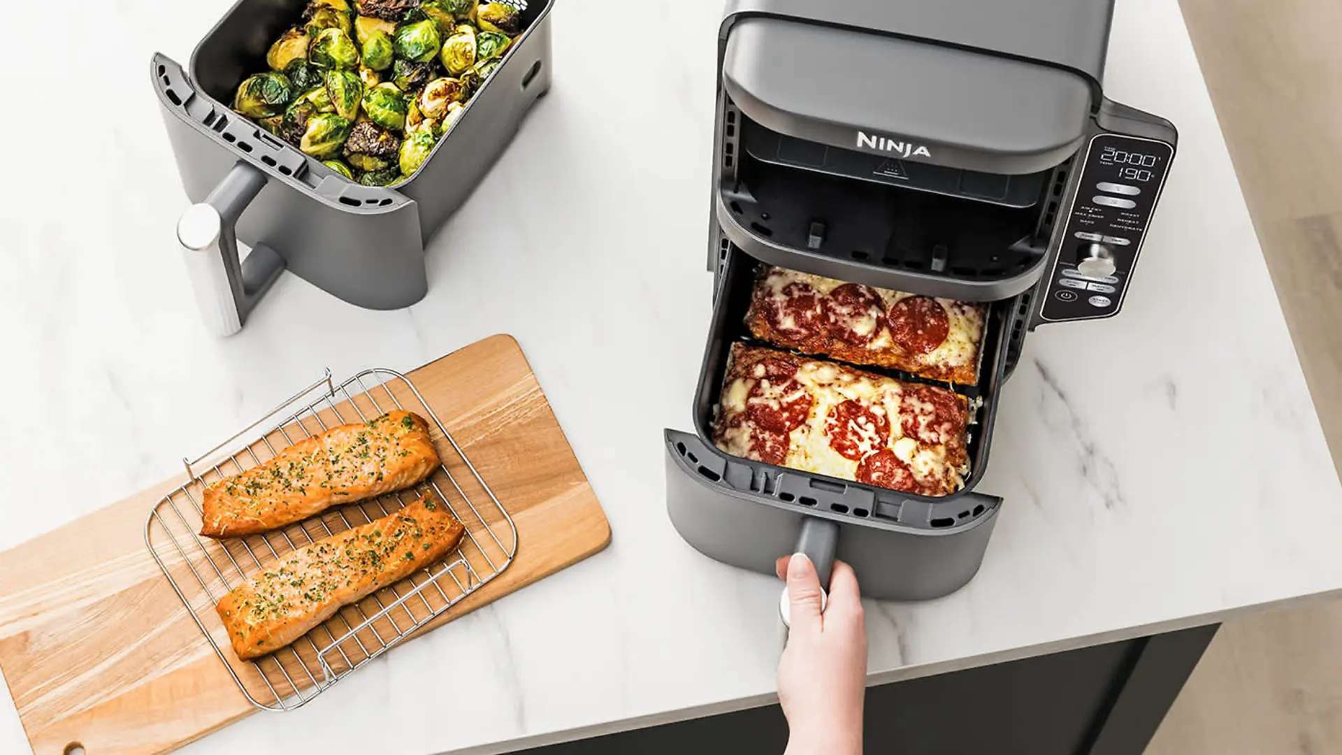 Heißluftfritteuse auf einer Arbeitsfläche, darin frsche Pizza. Daneben ein Rost mit Lachs und Rosenkohl.