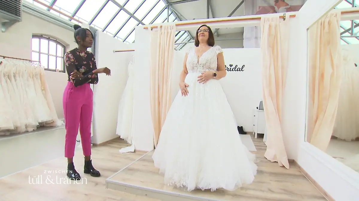 Zwischen Tüll und Tränen: Braut Ulli in einem ausladenden Brautkleid aus Tüll und Spitze vor dem Spiegel, daneben die Brautmodenexpertin