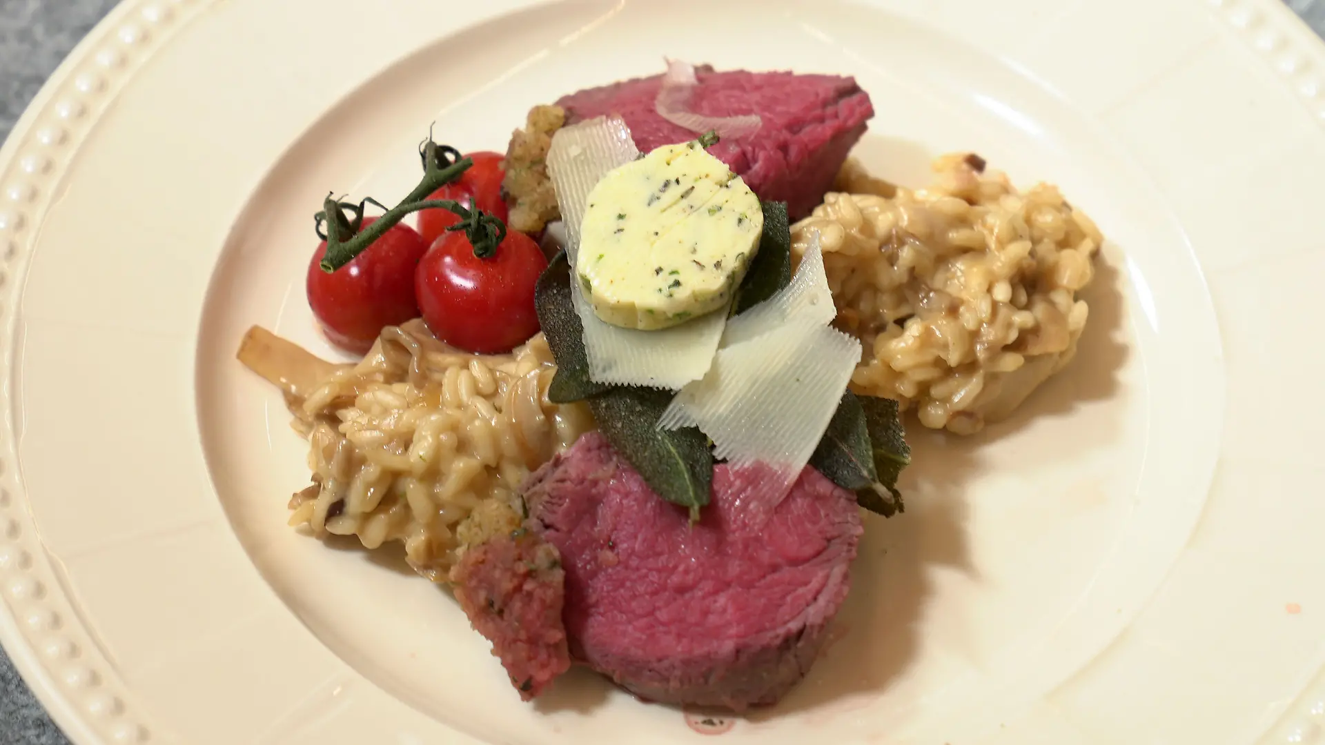 Das perfekte Dinner: Tellerbild: Rosa gegartes Fleisch an Risotto, dazu Kirschtomaten, Salbeiblätter, Parmesanhobel und Kräuterbutter