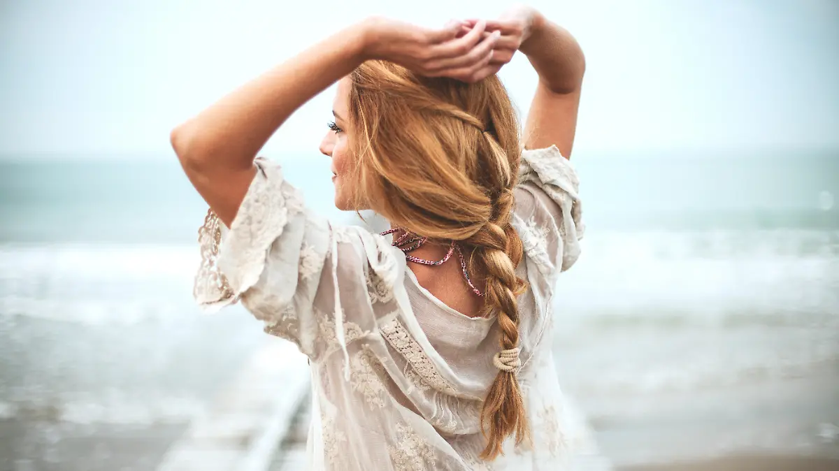 Frau in weißem Bohokleid, aufgenommen von hinten mit Strand und Meer im Hintergrund.