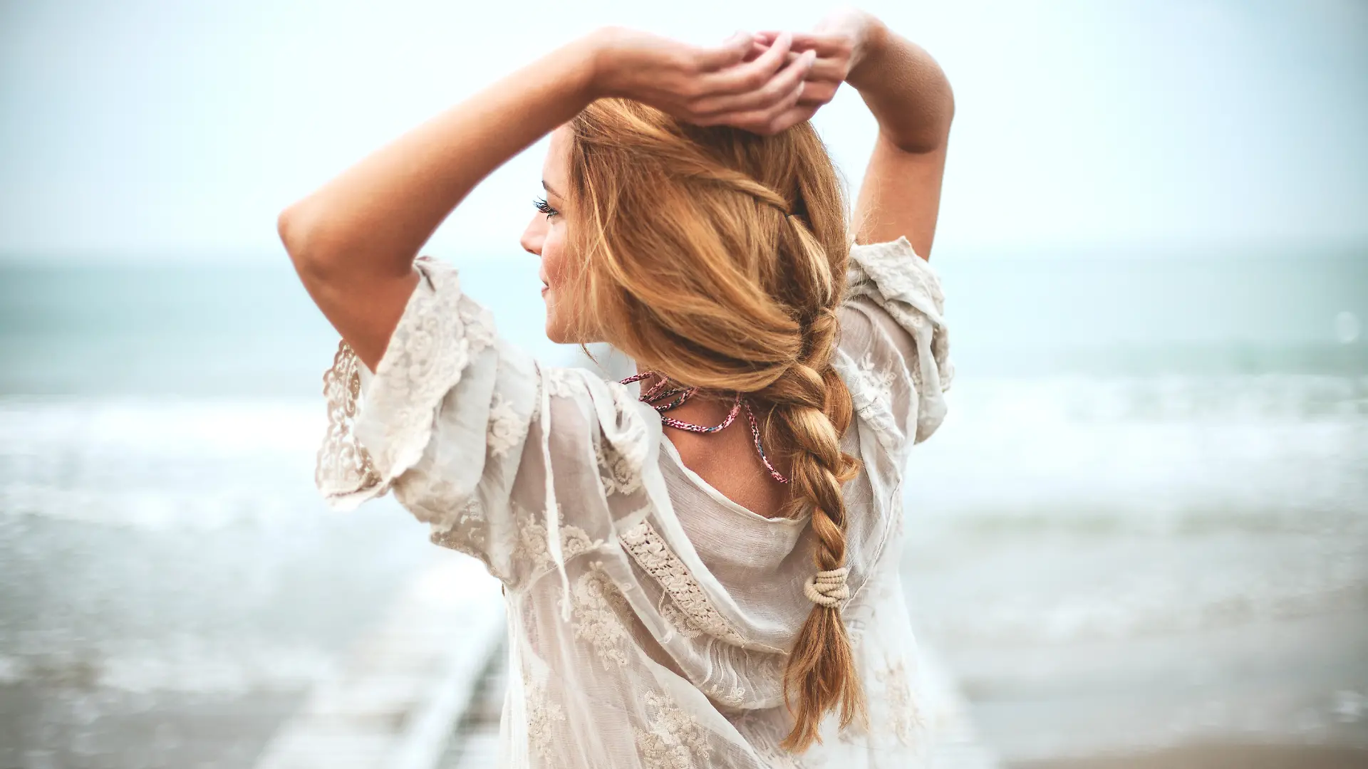 Frau in weißem Bohokleid, aufgenommen von hinten mit Strand und Meer im Hintergrund.