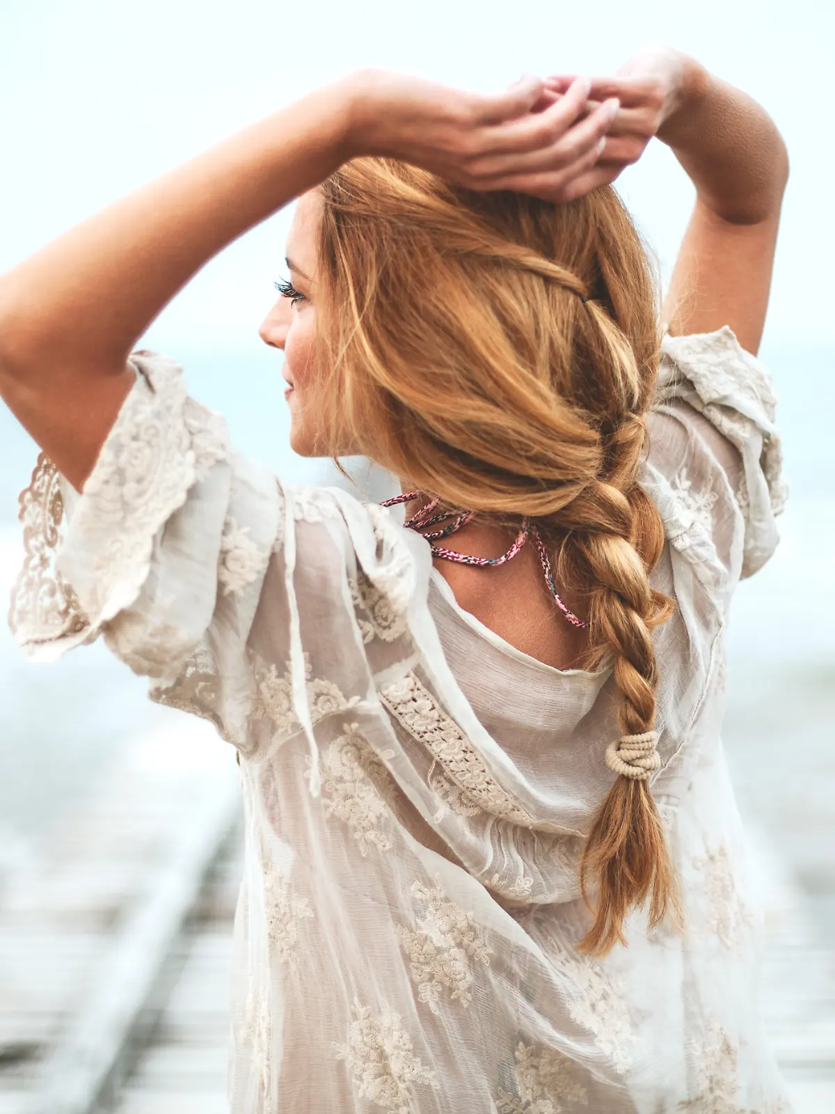 Frau in weißem Bohokleid, aufgenommen von hinten mit Strand und Meer im Hintergrund.