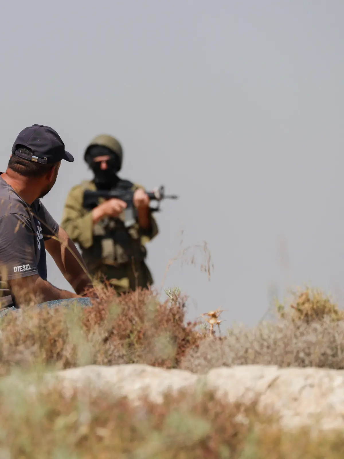 A Palestinian man sits on a rocky hillside as Israeli forces stand nearby amid smoke near Dura, southwest of Hebron, in the occupied West Bank, on June 9, 2026. Israeli forces dispersed Palestinians protesting against settlement activity near Hebron, where smoke and fires were seen during the confrontation.