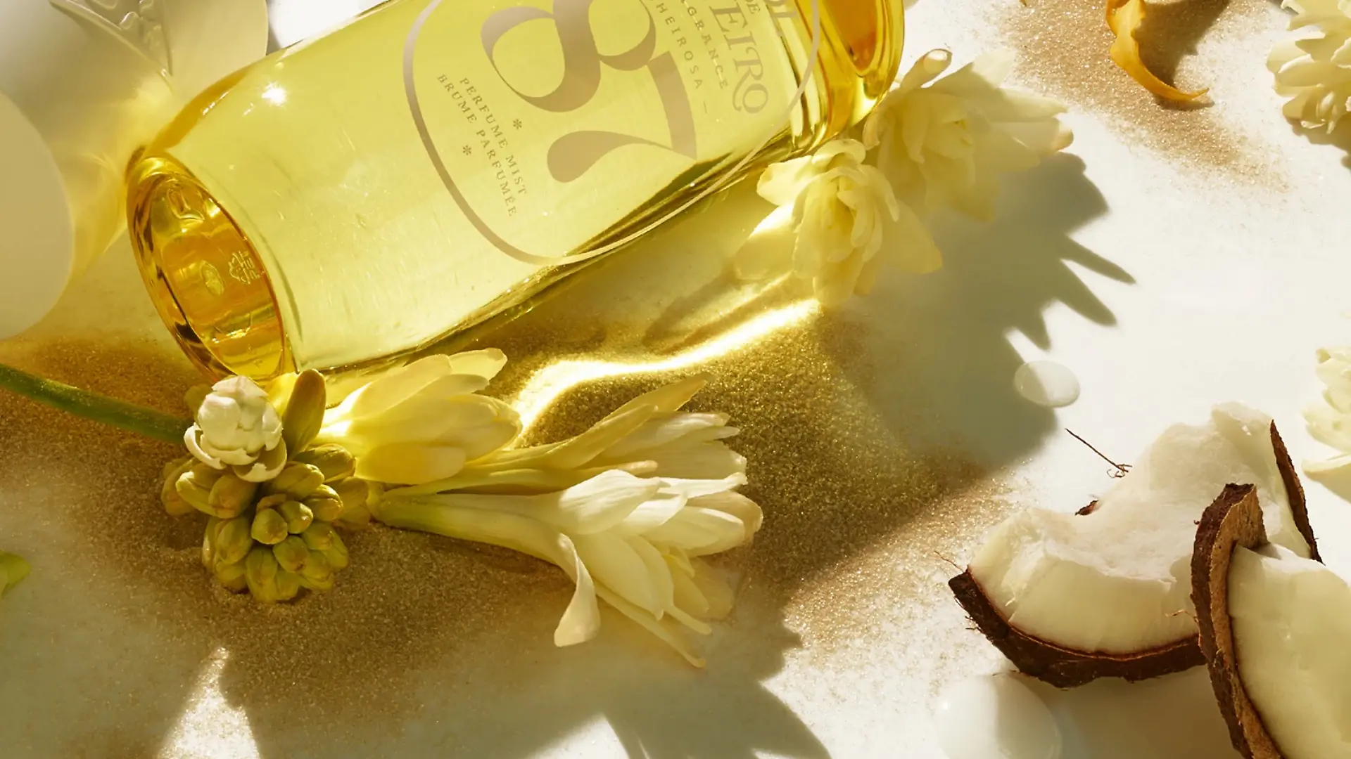 Das Parfum Sol de Janeiro Cheirosa ‘87 liegt mit Blumen und Kokosnuss auf einem Untergrund.