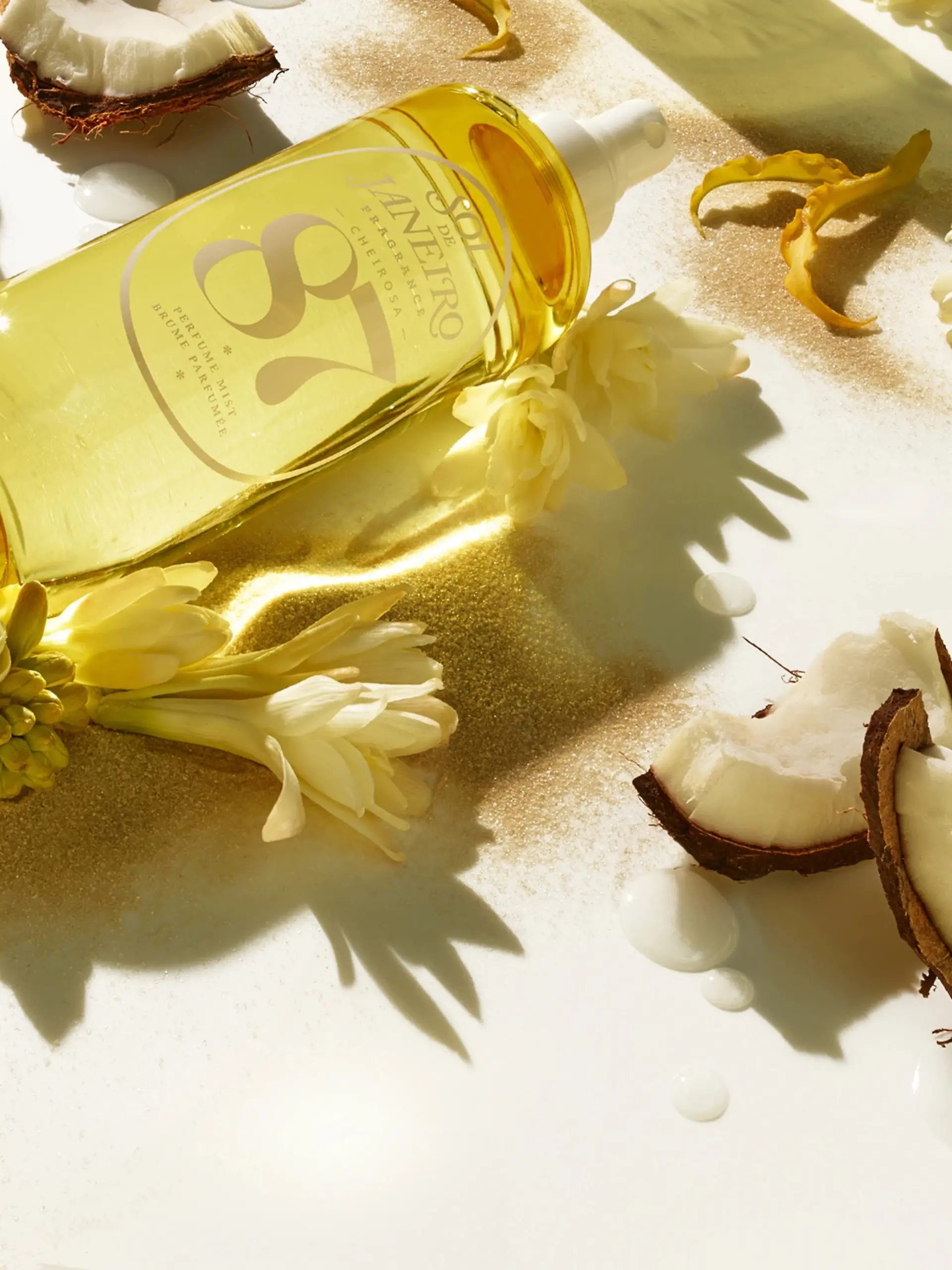 Das Parfum Sol de Janeiro Cheirosa ‘87 liegt mit Blumen und Kokosnuss auf einem Untergrund.