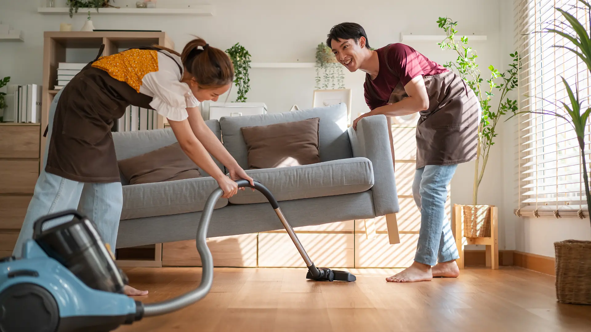 Zwei Menschen putzen zusammen ein modernes Wohnzimmer, ein Mann hebt die Couch an, die Frau saugt darunter.