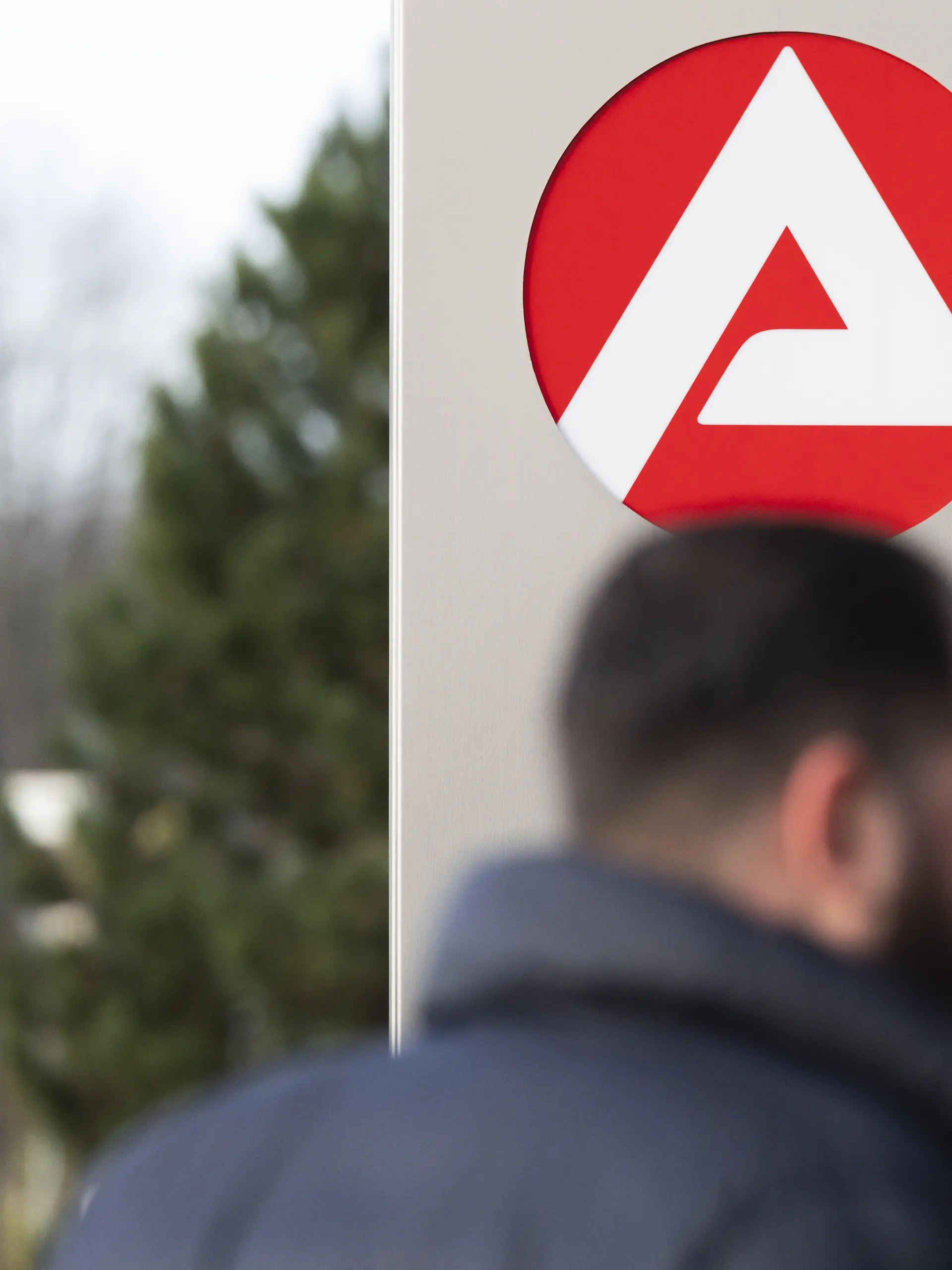 Stele mit dem Zeichen, Logo der Agentur für Arbeit vor dem Gebäude der Agentur für Arbeit, unscharfe Personen auf dem Weg zur Agentur, Köln, Nordrhein-Westfalen, Deutschland