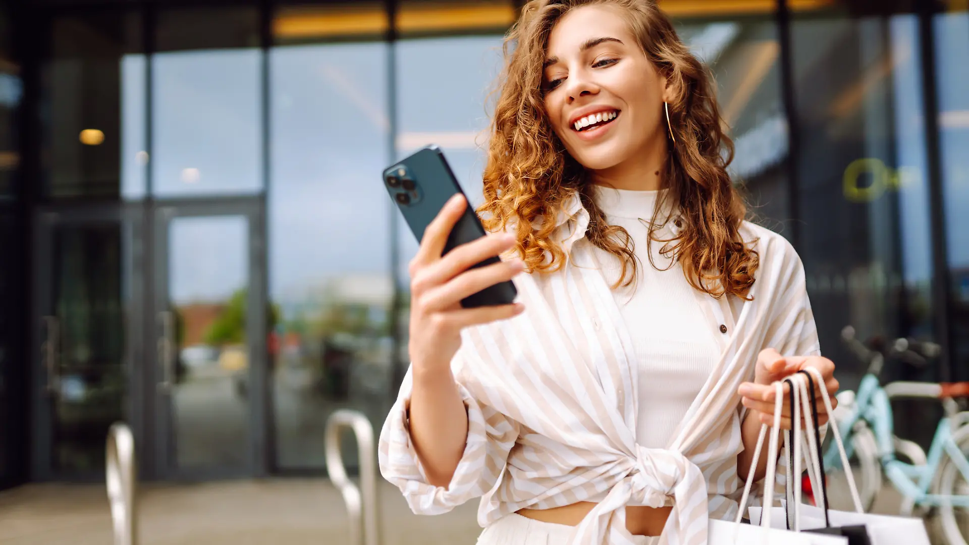 Eine Frau lacht und hat ein Smartphone und Einkauftaschen in der Hand.