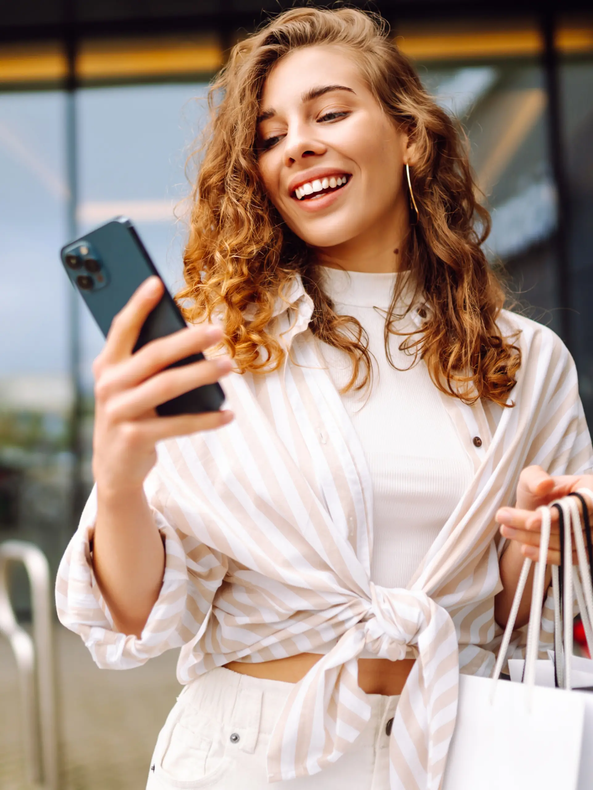 Eine Frau lacht und hat ein Smartphone und Einkauftaschen in der Hand.