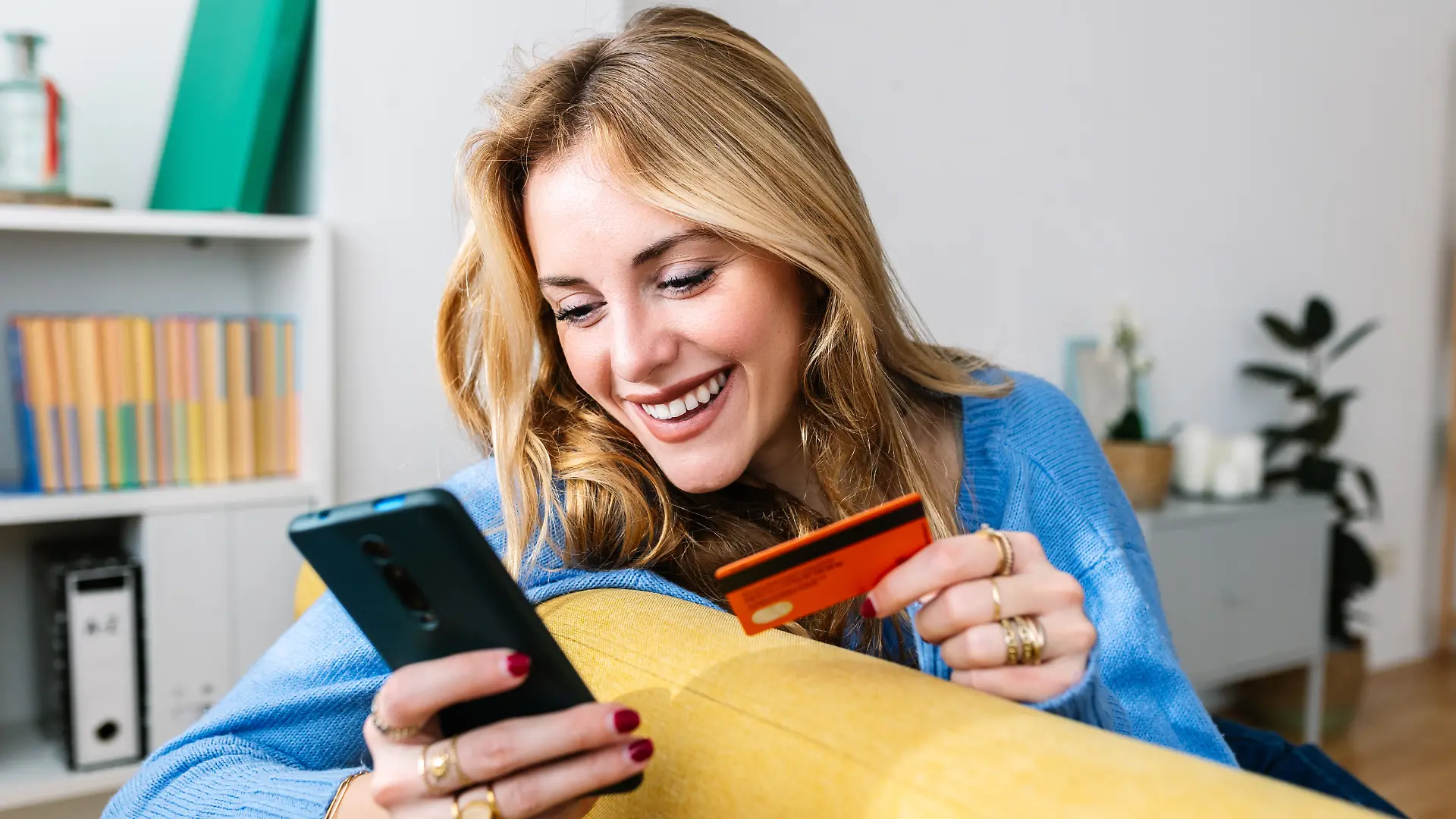 Eine fröhliche, junge, blonde Frau, die zu Hause beim Online-Shopping über eine Handy-App ihre Kreditkarte in der Hand hält. Konzept für E-Commerce und Lieferservice.