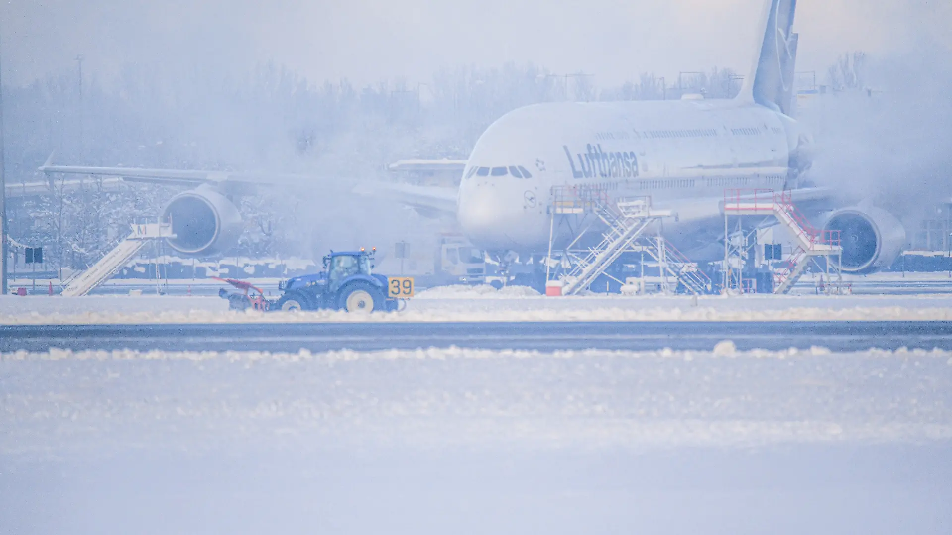 Ein Schneeräumer fährt im Schneetreiben auf dem Flughafen vor einer Maschine entlang. Nach einer mehrstündigen Unterbrechung wegen der Witterungsbedingungen hat der Münchner Flughafen seinen Betrieb wieder aufgenommen.