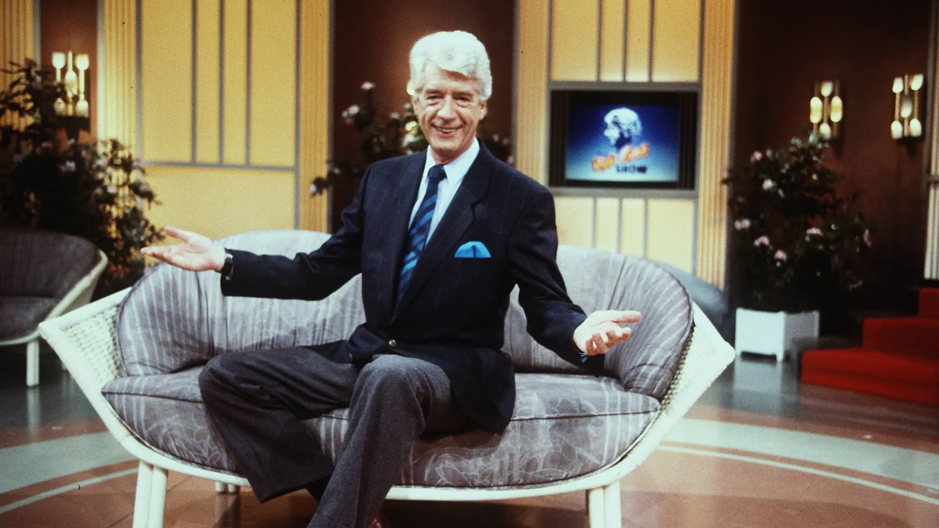 Rudi Carrell in Die Rudi Carrell Show Mann Moderation Entertainment Show Niederlande quer halb sitzend Kulisse Logo weißhaarig Anzug braun lächelnd gestikulierend