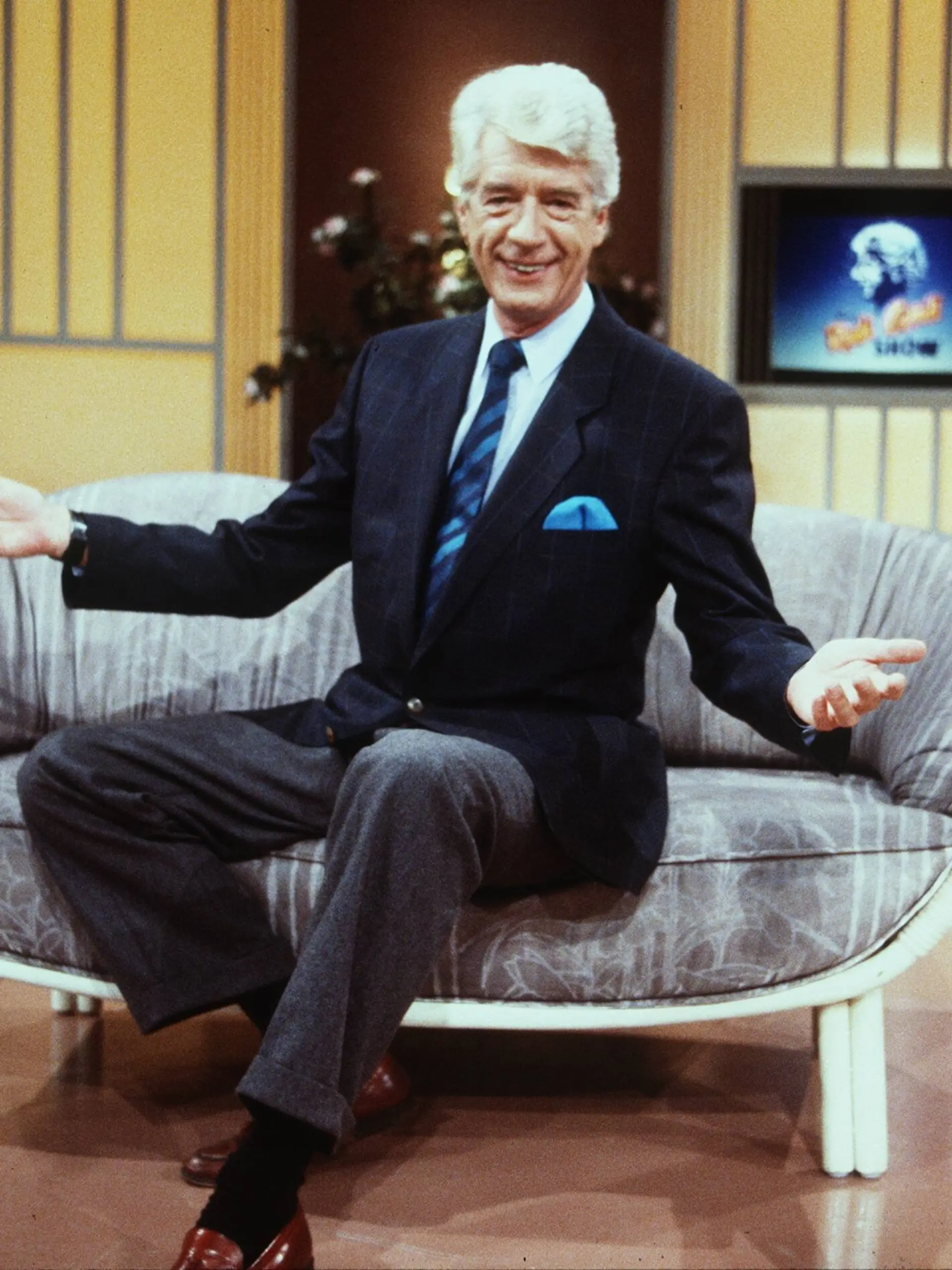 Rudi Carrell in Die Rudi Carrell Show Mann Moderation Entertainment Show Niederlande quer halb sitzend Kulisse Logo weißhaarig Anzug braun lächelnd gestikulierend
