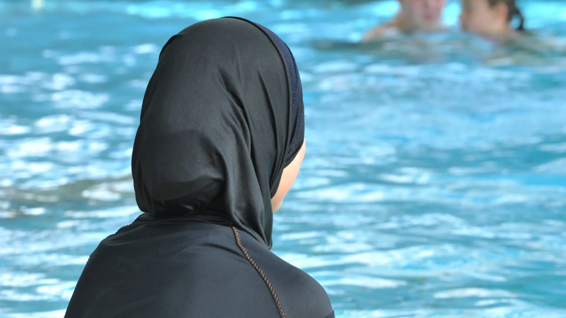 Eine Schülerin sitzt in einem Burkini am Rande eines Schwimmbeckens.