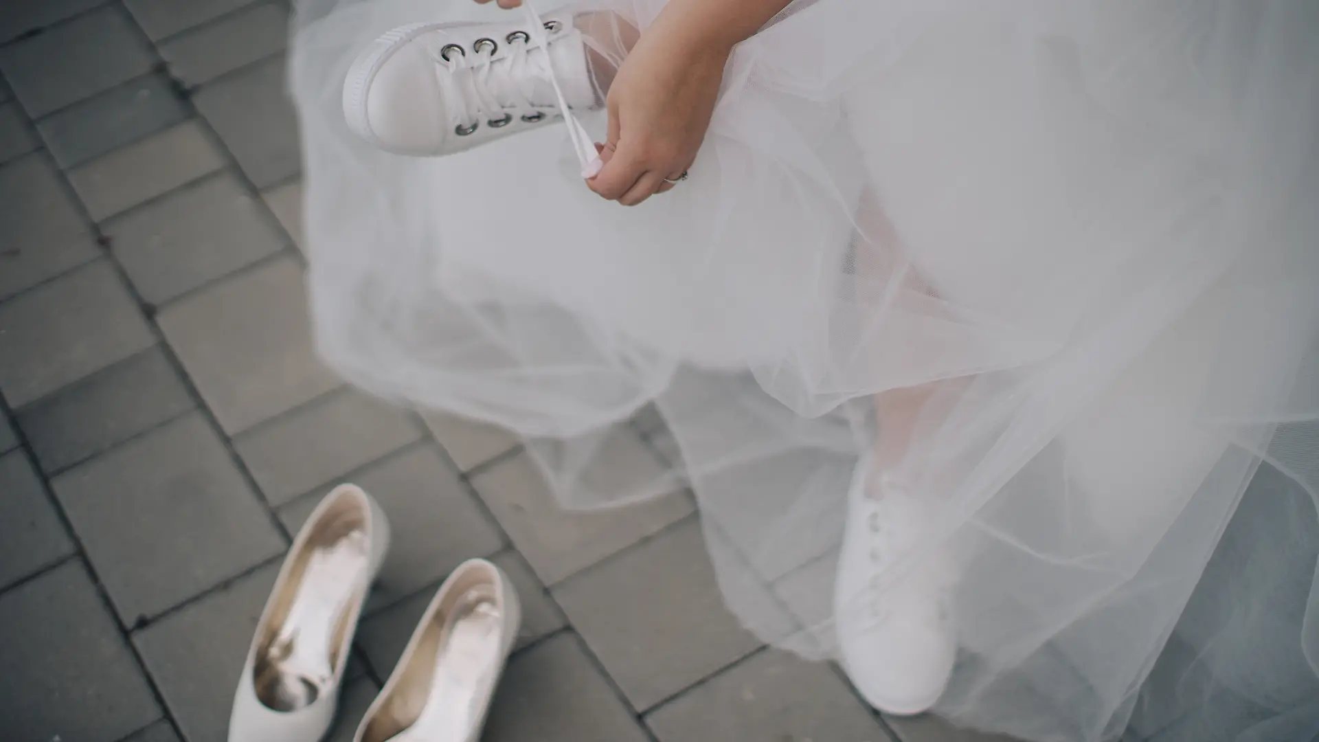 Frau im Hochzeitskleid zieht sich weiße Sneaker an, während ihre Absatzschuhe daneben stehen.