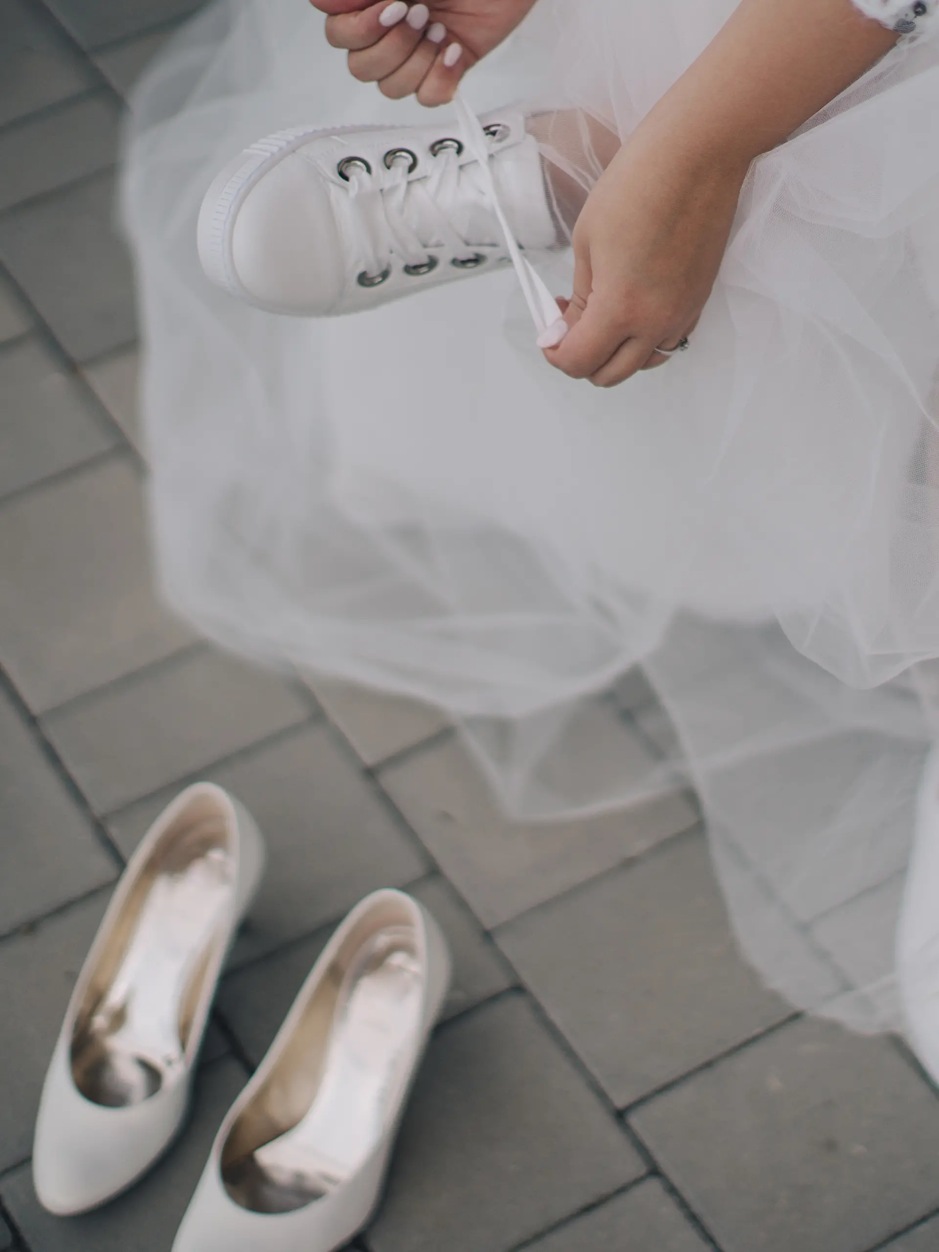 Frau im Hochzeitskleid zieht sich weiße Sneaker an, während ihre Absatzschuhe daneben stehen.